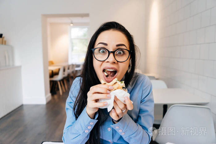 滑稽的年轻女孩在蓝色衬衣和杯子在工作以后在咖啡馆吃三明治