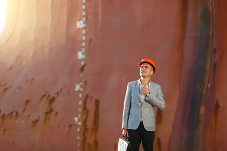 英俊的年轻胡子拉碴的成功商人灰色西装和保护性建筑橙色头盔持有的情况下, 站在海港对一艘生锈的船与水线背景