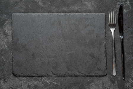 空黑色花岗岩石长方形板用叉子和刀子在黑纹理水泥背景, 顶面视图 vith 复制空间为您的文本