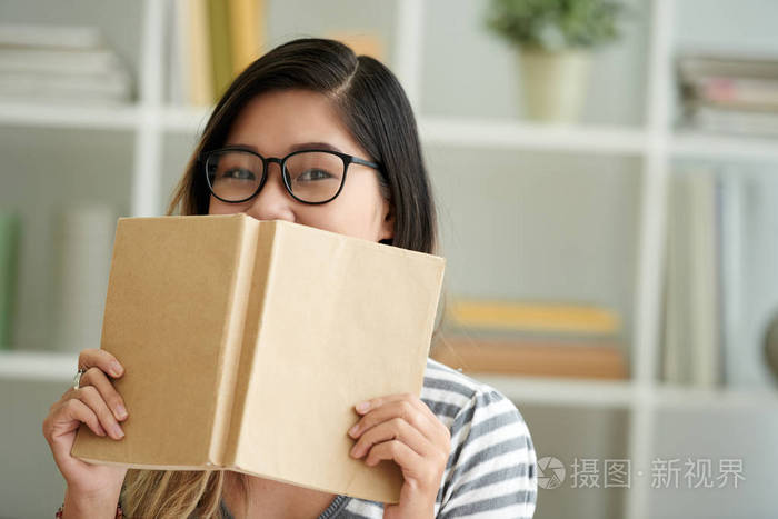 戴着一本书遮住脸的聪明女孩的肖像照片-正版商用图片0etewg-摄图新