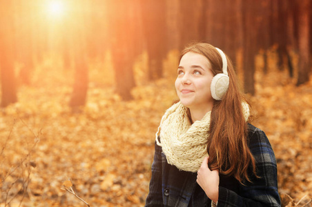 年轻美丽的女孩与温暖的围巾和耳罩仰望秋天的森林, 背景和阳光模糊