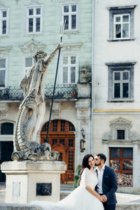 垂直婚礼肖像英俊的新郎轻轻地亲吻华丽的新娘在脸颊上, 而坐在市中心的老喷泉