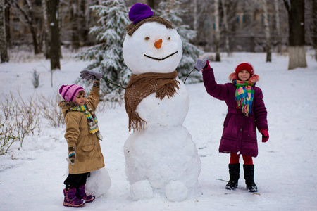 一个快乐的小女孩在大滑稽的雪人附近。一个可爱的小姐妹在冬季公园里玩得很开心, 冬天