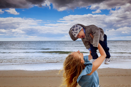 幸福的家庭。年轻的母亲吐到婴儿的天空，阳光灿烂的日子。肖像的母亲和小儿子在沙滩上