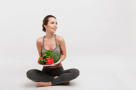 快乐的年轻女子坐在地板上与托盘的各种健康的蔬菜隔离在白色