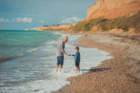 梦幻般的触摸场面英俊的父亲走在海边的石头海滩与可爱的儿子穿着时髦的衣服一起享受夏日时光