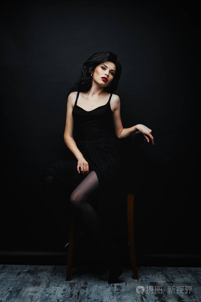华丽的年轻黑发妇女穿着优雅的黑色礼服, 对黑暗的工作室背景摆出