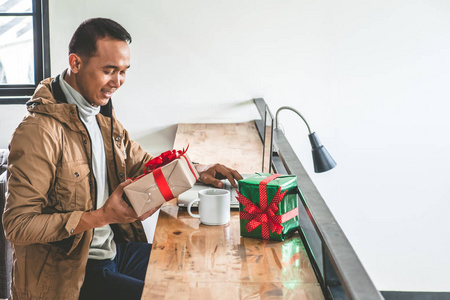 快乐的人手持礼品盒, 并在桌子上使用笔记本电脑。亚洲男子准备圣诞假期礼物