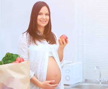 年轻的怀孕女子捧着一个苹果，站在厨房
