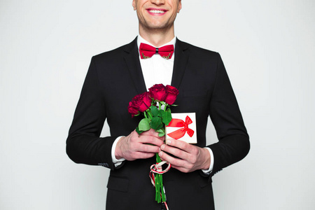 男子在正式的黑色西装和红色领结是手持礼品盒和美丽的红玫瑰在手中