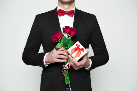 男子在正式的黑色西装和红色领结是手持礼品盒和美丽的红玫瑰在手中