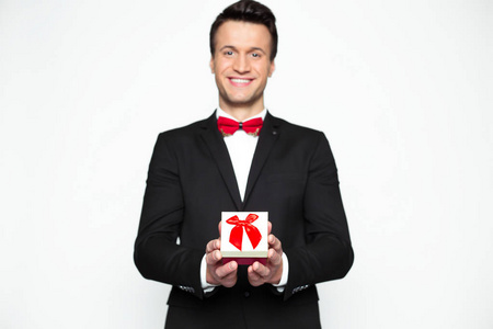节日礼物。在正式的黑色西装和红色弓领带的快乐英俊的现代男人拿着一个漂亮的礼品盒, 并显示在相机