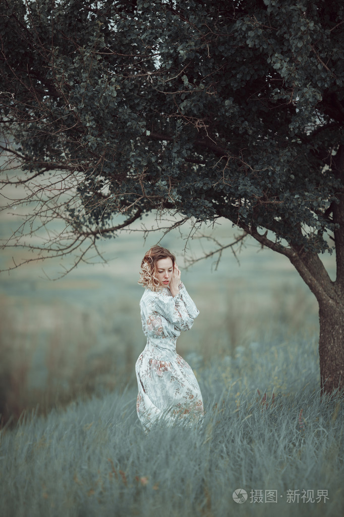 年轻的女孩穿着复古的服装站附近棵孤独的树