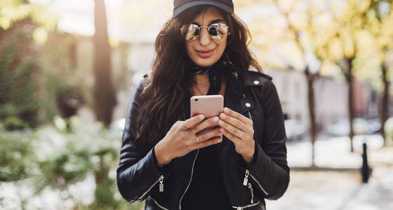 年轻女孩穿黑色皮夹克和使用现代智能手机室外