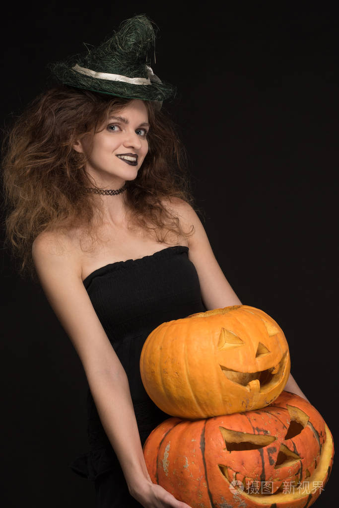 女巫,意味着与化妆漂亮女孩微笑着拿橘子南瓜在黑色背景上的万圣节