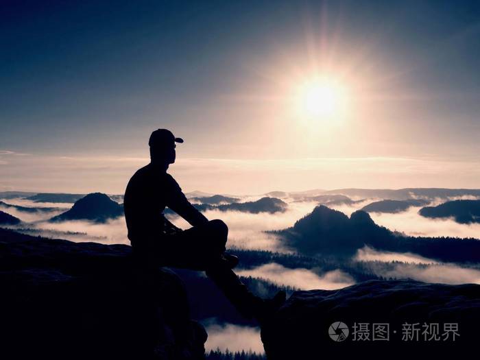 剪影的黑色和帽的独自徒步旅行者坐在悬崖上.蓝色滤光镜的照片.