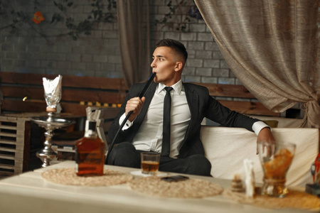 年轻时尚，穿着西装在酒吧休息。男人喝威士忌和抽烟的水烟