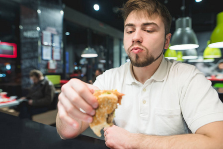 一个饥饿的人坐在快餐店里, 看着他手里的汉堡包。快餐概念