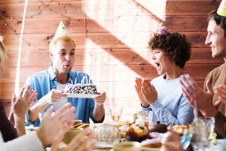 快乐的朋友祝贺年轻人在生日蛋糕上吹蜡烛的节日餐桌