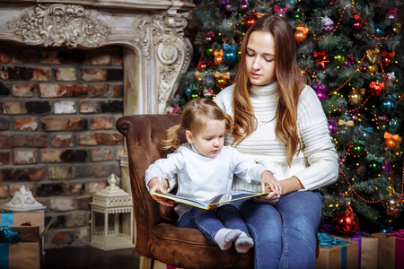 圣诞快乐，节日快乐 读一本书给她可爱的女儿，离室内圣诞树的年轻漂亮妈妈