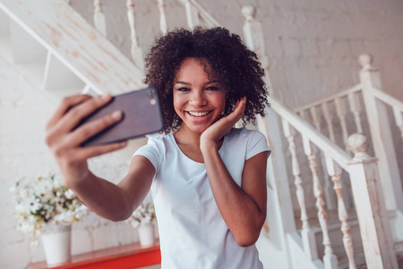 美丽的非洲女孩在白色的 t 恤让在家使用智能手机的拍照