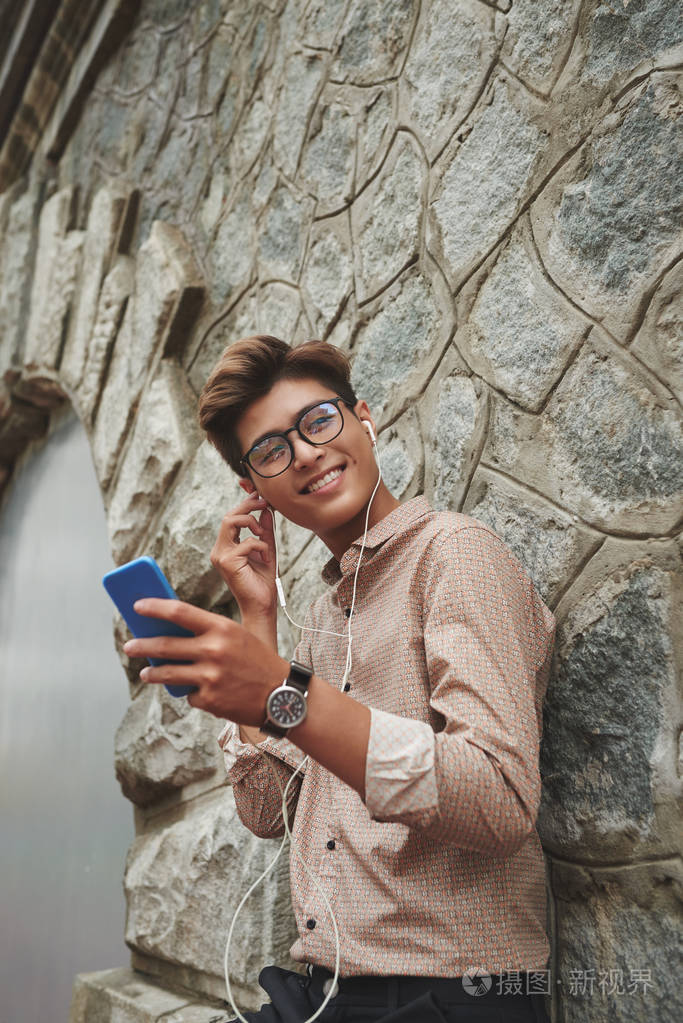 英俊的越南年轻人站在户外, 听音乐在他的智能手机