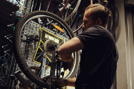 一个英俊的红头发的男性穿着牛仔裤套装, 在修理店里用自行车轮子工作。工人在车间里移除自行车轮胎