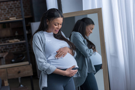 非洲裔美国人怀孕的妇女看着她的肚子, 而站在镜子