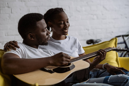 快乐的年轻非洲裔美国人夫妇坐在一起沙发和男子演奏声学吉他