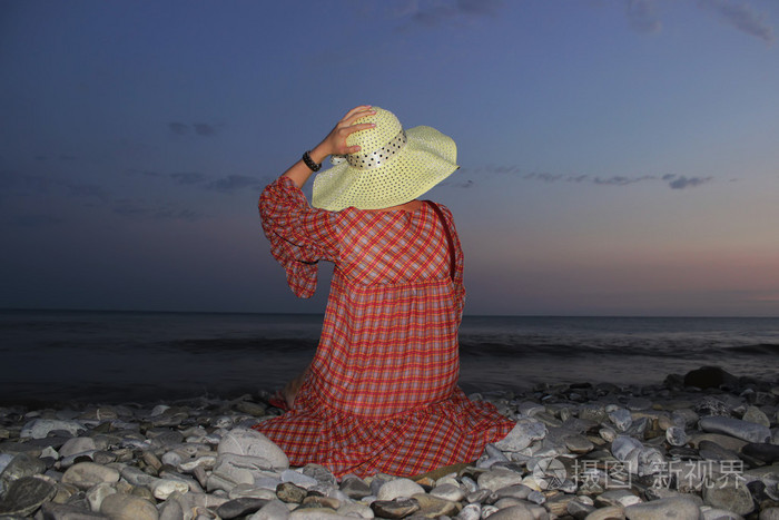 年轻漂亮的女孩坐在岸边的海在日落和抱着她用一只手的帽子