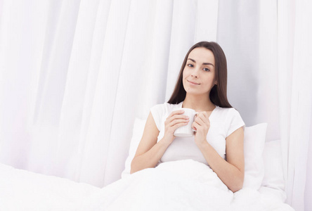 美丽年轻的黑发女孩喝着咖啡，微笑在白色的枕头上