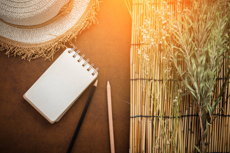 旅行概念以藤自然帽子与干燥花和树与空白页笔记本和铅笔在褐色皮革背景与轻的火光作用