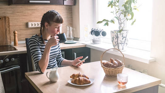 一个穿条纹衣服的女孩在家喝咖啡用羊角面包, 用智能手机照片
