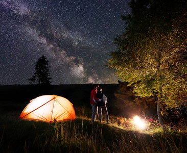 年轻情侣背包客站在火和树下的夜空与明亮的星星和银河在照亮橙色帐篷和山脉的背景下, 距离