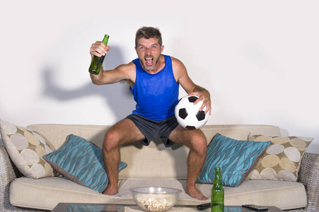 年轻有魅力的男人高兴和兴奋看足球比赛在电视上庆祝胜利目标跳在沙发沙发上, 啤酒和爆米花