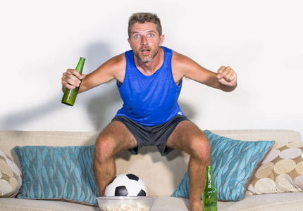 年轻有魅力的男人高兴和兴奋看足球比赛在电视上庆祝胜利目标跳在沙发沙发上, 啤酒和爆米花