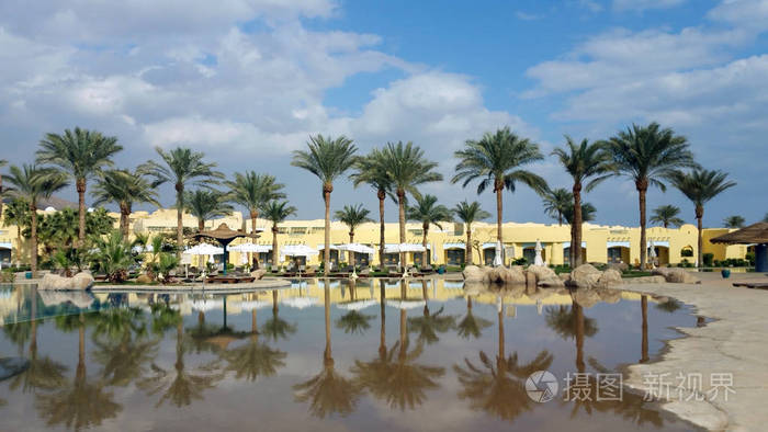 在埃及的度假胜地。棕榈和白色雨伞反射在水池水反对大厦和蓝天与白云
