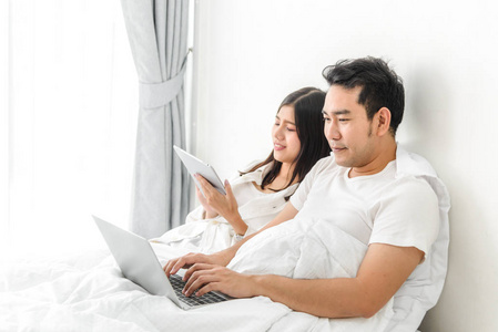 愉快的亚洲夫妇使用笔记本电脑和平板电脑一起在河上的床上
