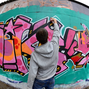 一个穿着灰色帽衫的年轻人在雨天的墙上画着粉红色和绿色的涂鸦。鱼眼射击