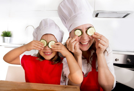 母亲和小女儿在厨师的帽子和一条围裙玩黄瓜片上厨房里的眼睛