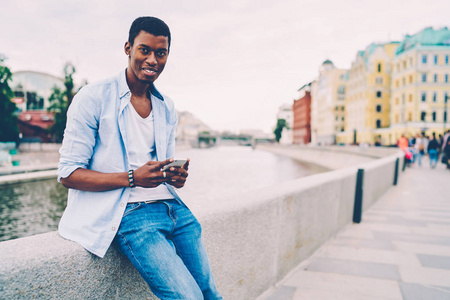 半长肖像微笑的美国黑人时髦的家伙发送信息通过电子邮件的智能手机在户外漫步。年轻积极的游客看着相机, 而花闲暇时间