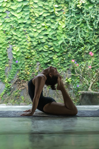 美丽的年轻女子的侧面观看练习瑜伽和表演背部弯曲与脚触摸头在墙面覆盖着绿叶