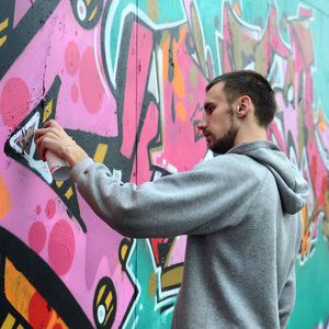 一个穿着灰色帽衫的年轻人在雨天的墙上画上了粉红色和绿色的涂鸦。