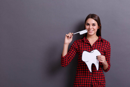 一个年轻快乐的女人拿着一张大牙齿的纸卡和一个灰色背景的牙刷。医学与牙医参观理念