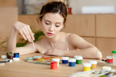 美丽的年轻女画家拿着刷子, 看着调色板在艺术工作室