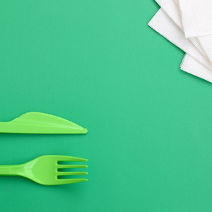 一次性塑料餐具绿色。塑料叉子和刀子躺在一个绿色的背景表面旁边的餐巾