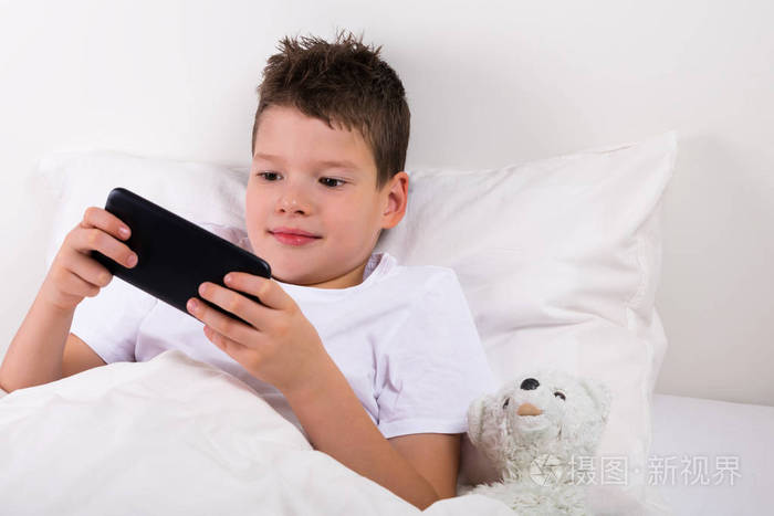 男孩玩游戏的电话躺在床上下一个玩具白色泰迪熊