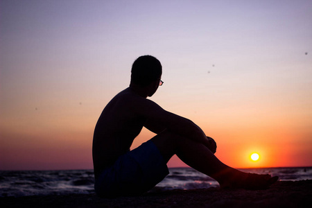 一个年轻的, 苗条的人反对夕阳背景的力量。一个男人坐在沙滩上, 看着海面上的日落。