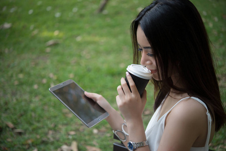 年轻美丽的女人持有一次性咖啡杯, 而短信通过智能手机在大自然户外