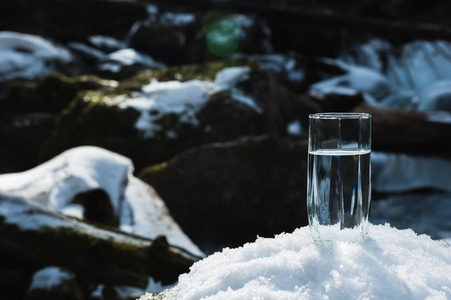 一个透明的玻璃杯, 饮用的山水在雪中矗立在冬日的一条清澈的寒山河的背景上
